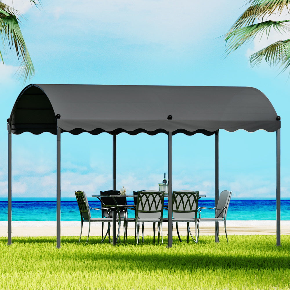 Long charcoal coloured gazebo or carport shading with outdoor table and chairs on the lawn with blue water beach background. 