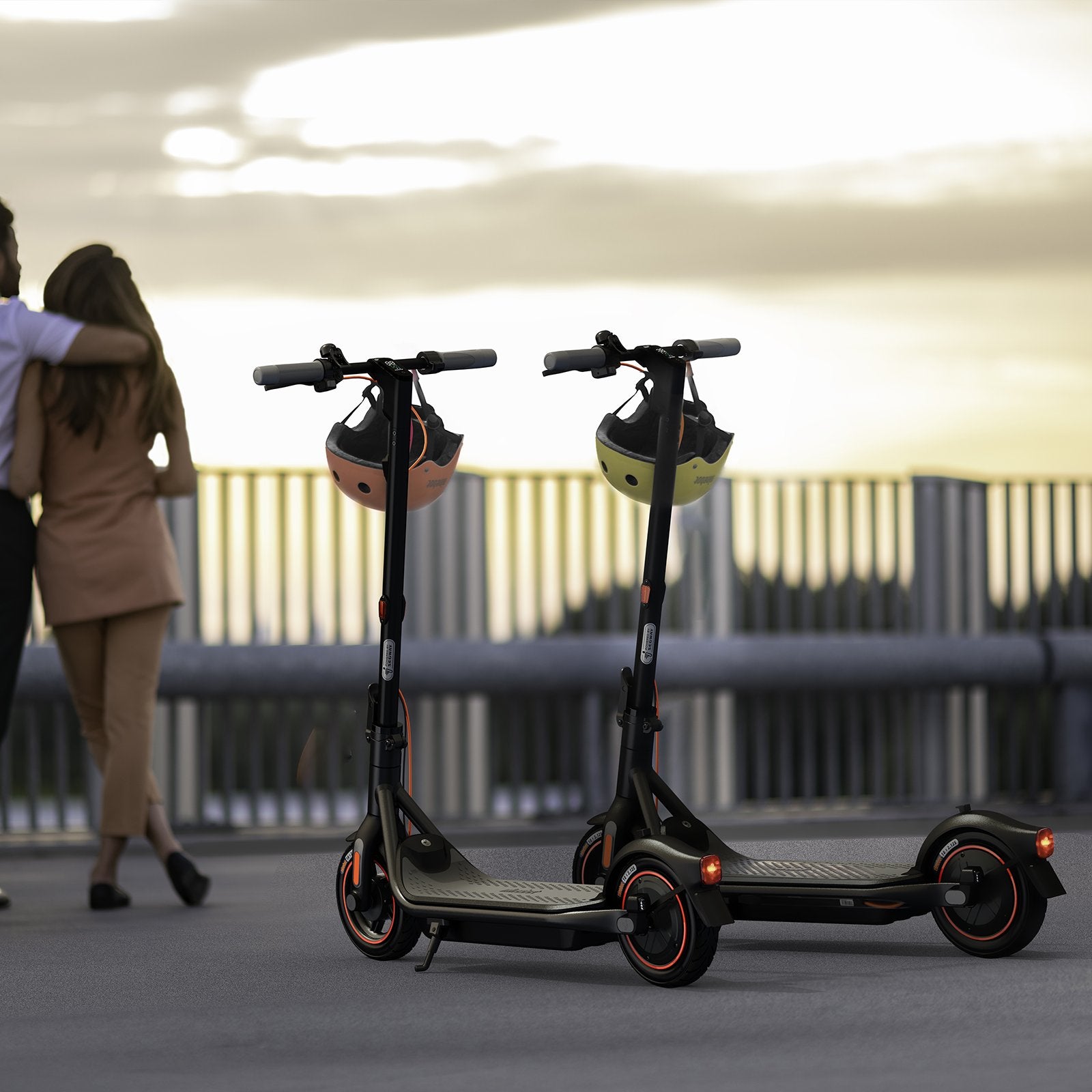 Two Segway Ninebot electric Scooter F65 in the foreground with a loving couple looking at a cloudy sunset from a bridge in the background.