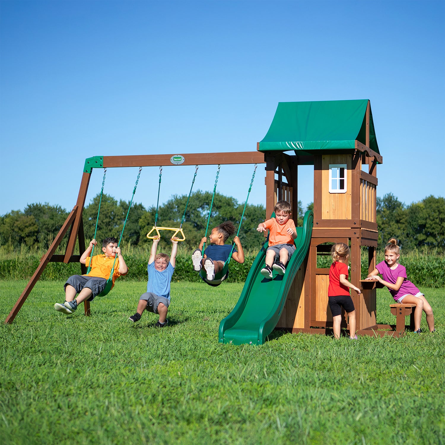 Six children playing on an outdoor play set with 3 swing a slide and cubby house with bench seat in a beautiful fresh green grassed area
